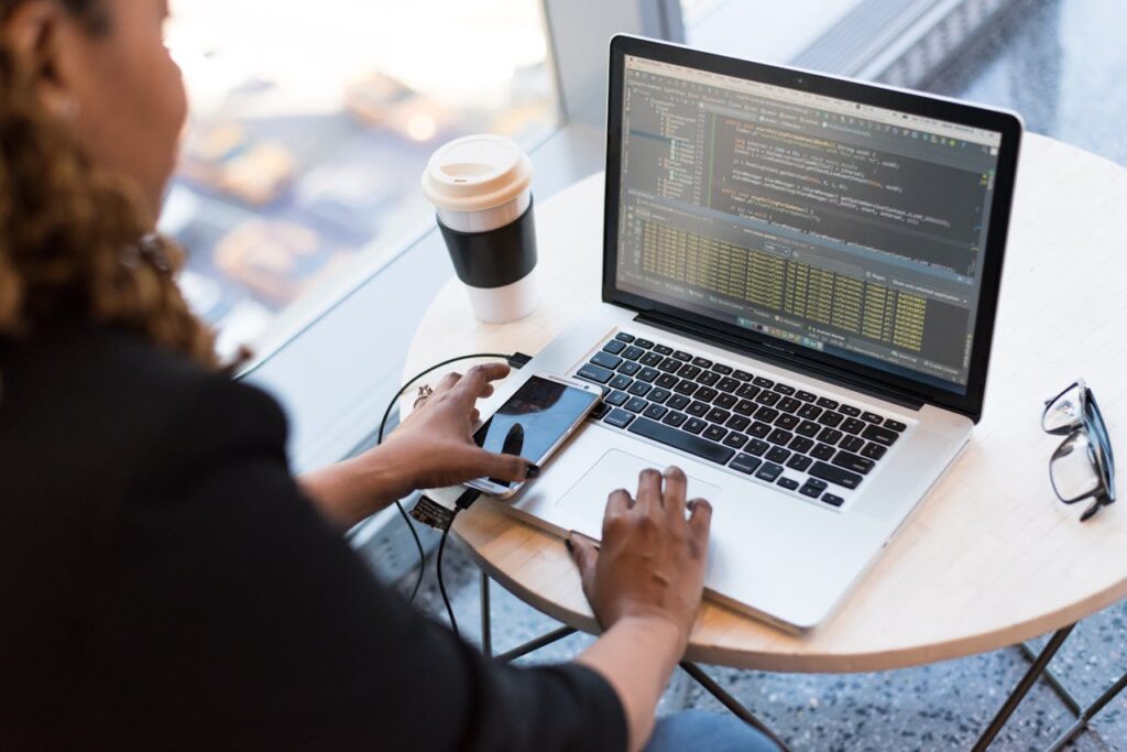 pexels photo 1181243 Black woman programming on a laptop with coffee, smartphone, and glasses on a desk in an office.